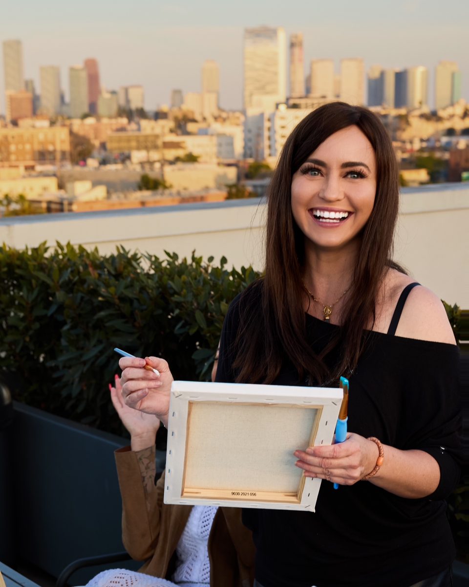 Zuzi Szokeova, founder of Wellness Atelier, smiling while holding a blank canvas at a rooftop painting event in Los Angeles